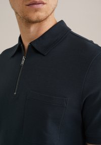 Polo de color azul marino hecho de tejido suave, con cuello con cremallera y un bolsillo en el pecho, con un diseño ajustado y textura mínima.