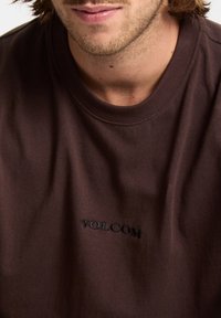 T-shirt en coton marron avec un col rond et un logo "VOLCOM" brodé en noir sur le devant. Le tissu a une texture lisse.