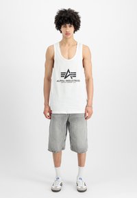 Mladenič s kodrastimi lasmi, oblečen v bel brezrokavnik, sivi hlačnici do kolen, belimi nogavicami in črno-belimi supergami, stoji.