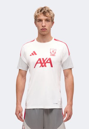Mladi moški s kratkimi blond lasmi, oblečen v belo-rdečo dres Liverpool Football Club in sive kratke hlače na preprostem ozadju.