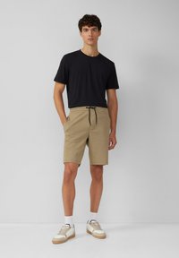 T-shirt noir, shorts beige avec cordon de serrage, et chaussettes blanches avec baskets beiges. Tissu lisse, coupe décontractée et style casual.
