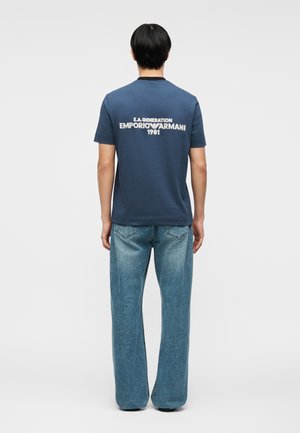 Homme debout de dos, portant un t-shirt bleu Emporio Armani avec texte blanc et un jean bleu ample sur un fond uni.