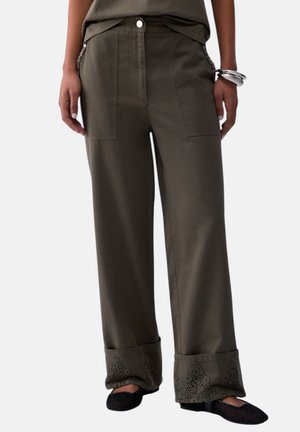 Donna che indossa pantaloni a gamba larga verde oliva con dettagli in pizzo sull'orlo risvoltato e scarpe basse nere, mani rilassate ai lati.