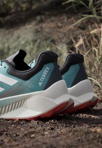 Paar türkisfarbene und schwarze Outdoor-Laufschuhe mit weißen Sohlen, die auf kiesigem Boden in natürlicher Außenumgebung ruhen.