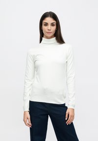 Λευκό πουλόβερ με ζιβάγκο από ελαφρύ ύφασμα, με μακριά μανίκια και εφαρμοστή γραμμή, συνδυασμένο με σκούρο μπλε τζιν παντελόνι.