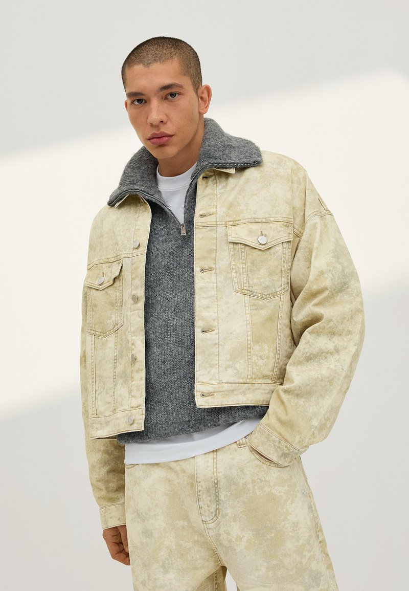 Mann mit kurzen Haaren, der eine ausgebleichte beige Jeansjacke und Hose trägt, darüber einen grauen Reißverschluss-Pullover und ein weißes Hemd, stehend vor einem hellen Hintergrund.