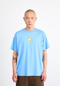 Light blue cotton t-shirt featuring a small yellow square logo with black and white stripes at the chest. Classic crew neck and short sleeves.