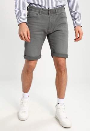 Homme portant un short en denim gris retroussé, des baskets blanches montantes, des chaussettes blanches et une chemise à manches longues rayée, debout devant un fond uni.