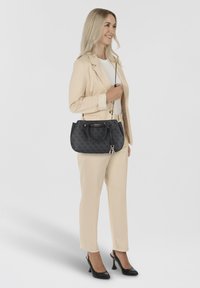 Sac à main noir avec un motif subtil, accents en doré, et deux poignées, tenu par une personne en costume beige clair et talons noirs.