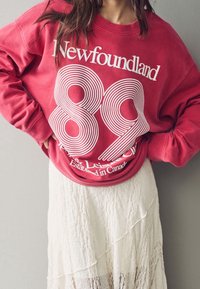 Felpa rossa con tipografia retro bianca che recita "Newfoundland 89" e "Established in Canada", abbinata a una gonna bianca e testurizzata.
