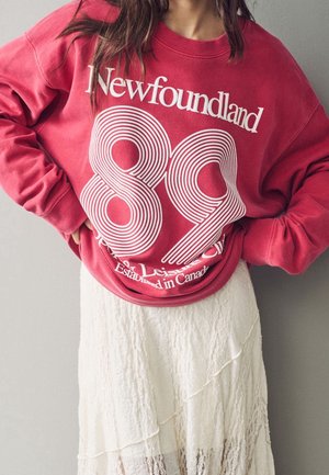 Sweat-shirt rouge avec une typographie rétro blanche portant "Terre-Neuve 89" et "Établi au Canada", associé à une jupe blanche texturée.