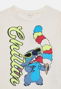 Hvit T-skjorte med en tegneseriefigur i solbriller, blå pels, som holder en fargerik iskjeks. Tekst "Chillin" i lys gul.