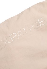 Beige stof med hævet, blank, prægede tekst, der staver "EXPATRIE." Let tekstur med en mat baggrund. Minimalistisk design.