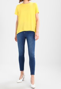 Žlutý krátký rukáv s rovný střihem, v kombinaci s tmavě modrými skinny džíny. Bílé vysoké podpatky dotvářejí celkový vzhled.