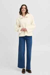 Kravefarvet fleecepullover med lynlåskrave, brede ærmer og struktureret overflade, parret med blå brede denim jeans og brune sko.