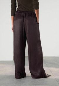Ciemnofioletowe spodnie o szerokich nogawkach wykonane z gładkiego satynowego materiału, wyposażone w elastyczny pas i boczne kieszenie, pokazane od tyłu.