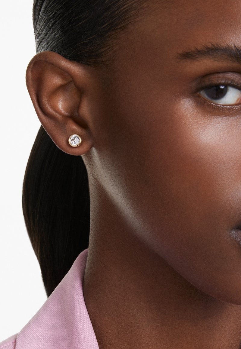 Gull øredob med en klar rund krystall i midten, vist på et nært bilde av et øre. Hudtonen er mørk med en glatt tekstur.