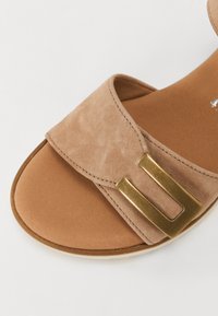 Beige suede sandal with a wide strap featuring a gold-tone metal buckle. The textured upper contrasts with a smooth, light brown insole.