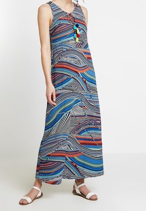 Abito maxi senza maniche con righe ondulate colorate in blu, rosso e nero, scollo annodato con nappine multicolore, indossato con sandali bianchi.