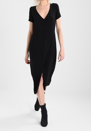 Žena v černých midi šatech s výstřihem do V a krátkými rukávy, se zapínáním na knoflíky vpředu, v kombinaci s černými kotníkovými botami, kráčející vpřed.