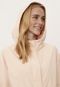 Vaaleanpunainen huppujakku, jossa on vetoketjukiinnitys, rento malli ja hienovaraiset ompelupisteet. Kangas on sileä ja matta.