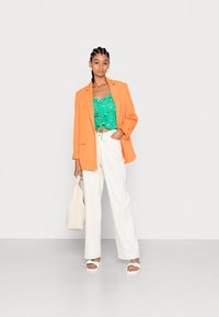 Blazer laranja, top de flores verde, calças largas brancas e sandálias brancas. Segura uma bolsa bege. Tecidos suaves e ajuste casual.