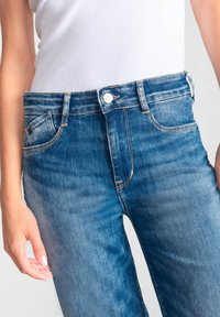 Jean en denim bleu taille haute avec un délavage, cinq poches, fermeture à bouton et accent en fil doré visible.