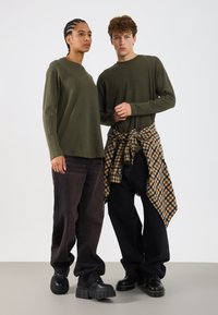 Divi pieaugušie, tērpušies olīvzaļos garajās piedurknēs t-kreklos un melnās biksēs, no kuriem viens valkā kreklu ar rakstu, sasietu ap vidukli, stāv vienkāršā fonā.