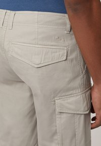 Bež cargo hlače s glatkom teksturom, dva zadnja džepa i jedan bočni džep. Imaju prošivene detalje i mali logo iznad desnog džepa.