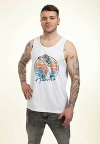 Hvid tanktop med en farverig grafik af en karakter og "Hakuna" tekst. Båret med grå slidte jeans, der viser tatoveringer på armene.