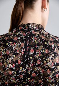 Fekete virágos csipke felső, kis rózsákkal és százszorszépekkel díszítve, magas nyakkal és finom, fodros szegéllyel, hátsó cipzárral zárva.