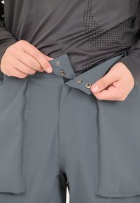 Grijze broek met een gladde textuur, voorzien van een drukknoop- en ritssluiting. Een persoon bevestigt de taille met één hand.
