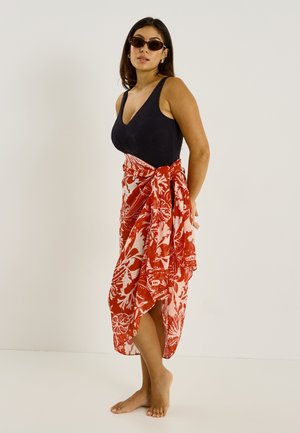 Mujer con bañador tipo tankini negro y sarong floral rojo y blanco anudado en la cintura, con gafas de sol oscuras, de pie descalza.