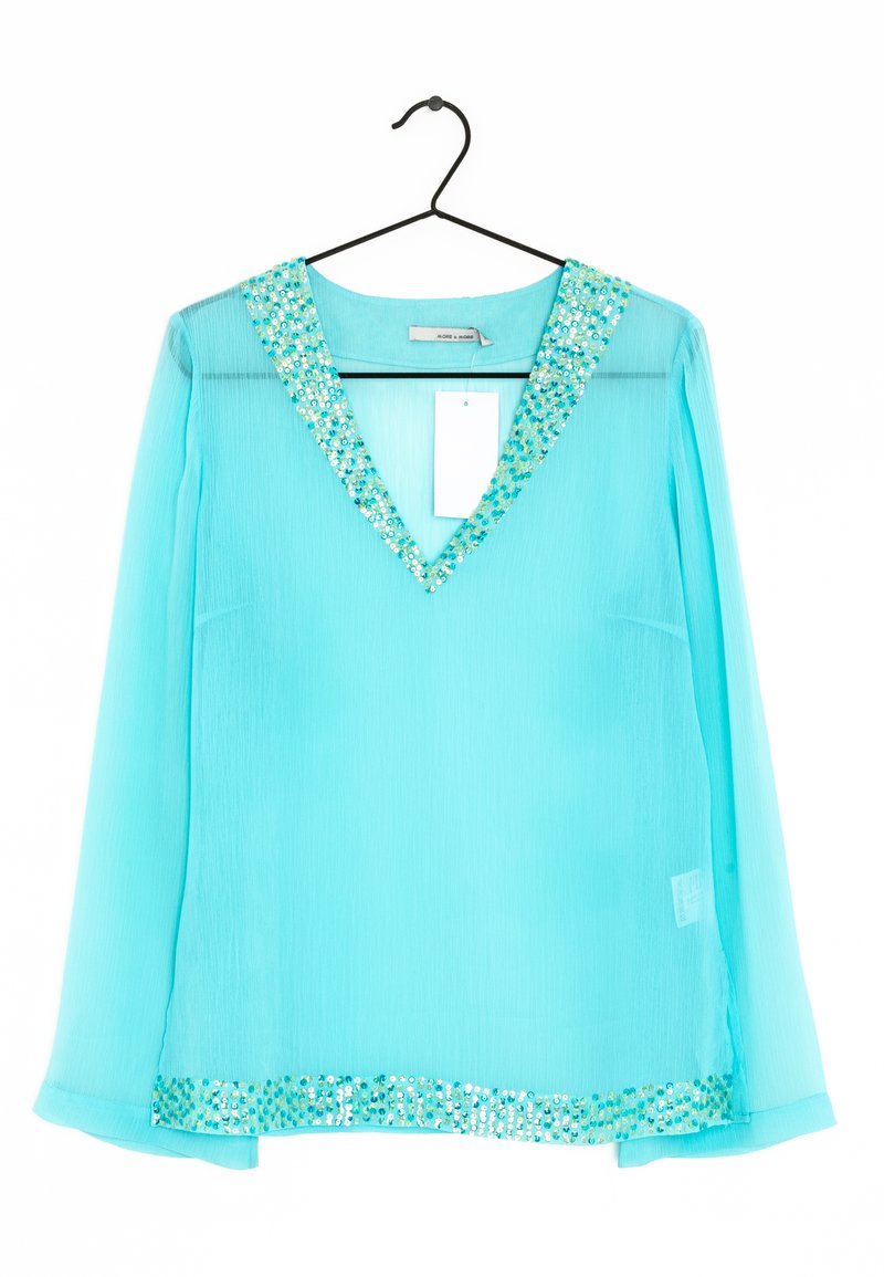 Turkis bluse med transparent stof, V-udskæring prydet med runde pailletter, lange ærmer og en pailletkant.