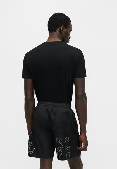 Άνδρας με την πλάτη, φοράει απλό μαύρο t-shirt και μαύρα σορτς με λευκά λογότυπα βελών και κείμενο σε ανοιχτό γκρι φόντο.