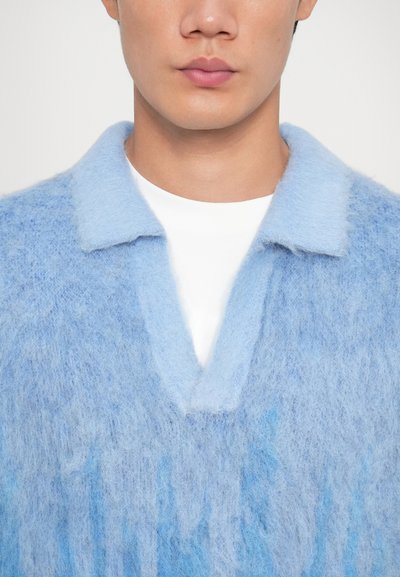 Maglione a maglia pelosa blu chiaro con colletto e apertura a V. Texture morbida con sottili variazioni di tonalità. Maglietta bianca semplice visibile.