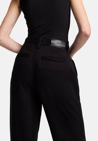 Pantalon noir à coupe droite, avec poches arrière et une étiquette à la taille. Le matériau semble solide avec une texture lisse.