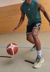 Camiseta sin mangas verde, pantalones cortos negros con acentos blancos, jugando al baloncesto. Lleva zapatillas verde claro, calcetines azules y está botando un balón rojo.