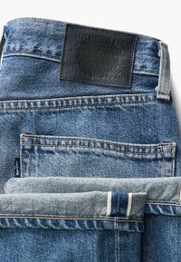 Jeans de mezclilla azul con un parche de cuero negro. Presentan un diseño clásico de cinco bolsillos y costuras expuestas. El dobladillo doblado revela una tela a rayas.
