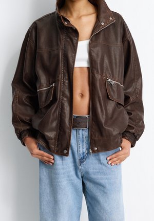 Personne portant une veste en cuir marron foncé sur un crop top blanc et un jean bleu clair avec une ceinture noire, debout les mains dans les poches.