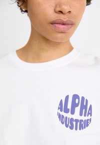 Balts kokvilnas T-krekls ar lielu apļveida grafiku violetā krāsā, kurā rakstīts "ALPHA INDUSTRIES". Vienkāršs, ar apaļu kakla izgriezumu.