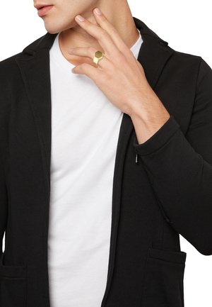 Złoty heksagonalny pierścień na dłoni, na której znajduje się czarna kurtka z długim rękawem na białym T-shircie. Uwaga skupiona na wzorze pierścienia i fakturze tkaniny.