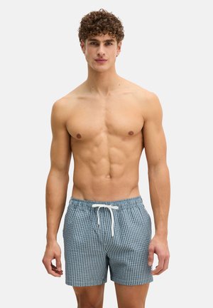 Jonge man met krullend haar, gekleed in blauw-witte gestreepte zwemshorts met een witte trekkoord, staand tegen een effen achtergrond.