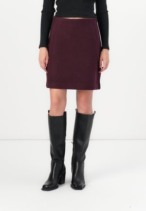 Mujer con falda ciruela hasta la rodilla, top negro de manga larga y botas negras de cuero con tacón hasta la rodilla, de pie sobre un fondo blanco.