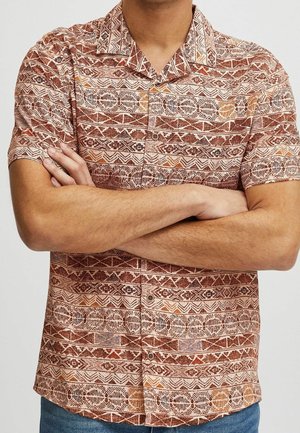 Homme portant une chemise à manches courtes avec des motifs géométriques orange et marron, bras croisés, et jean bleu.