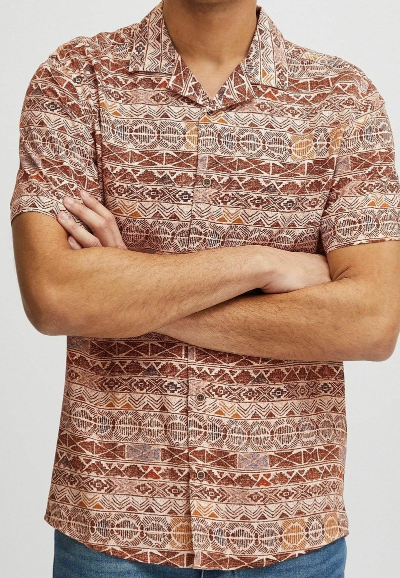 Homme portant une chemise à manches courtes avec des motifs géométriques orange et marron, bras croisés, et jean bleu.