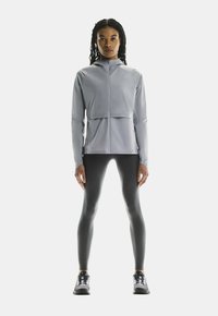 Veste zippée gris clair avec un design court, associée à des leggings gris foncé et des chaussures de sport noires. Tissu lisse, style minimaliste.