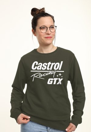 Frau mit Brille und Hochsteckfrisur, trägt ein olivgrünes Sweatshirt mit weißem "Castrol Racing GTX"-Schriftzug und hellblaue Jeans.