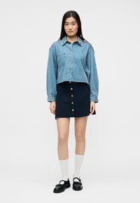 Femme portant une chemise en denim bleu avec un col orné, une jupe noire boutonnée, des chaussettes blanches et des chaussures Mary Jane noires, debout devant un fond blanc.