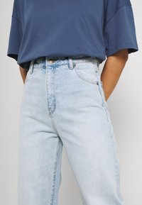 Jeans de ganga azul claro, de cintura alta, com um corte reto, apresentando bolsos frontais, fechos em botão dourado e detalhes de desvanecimento subtil.